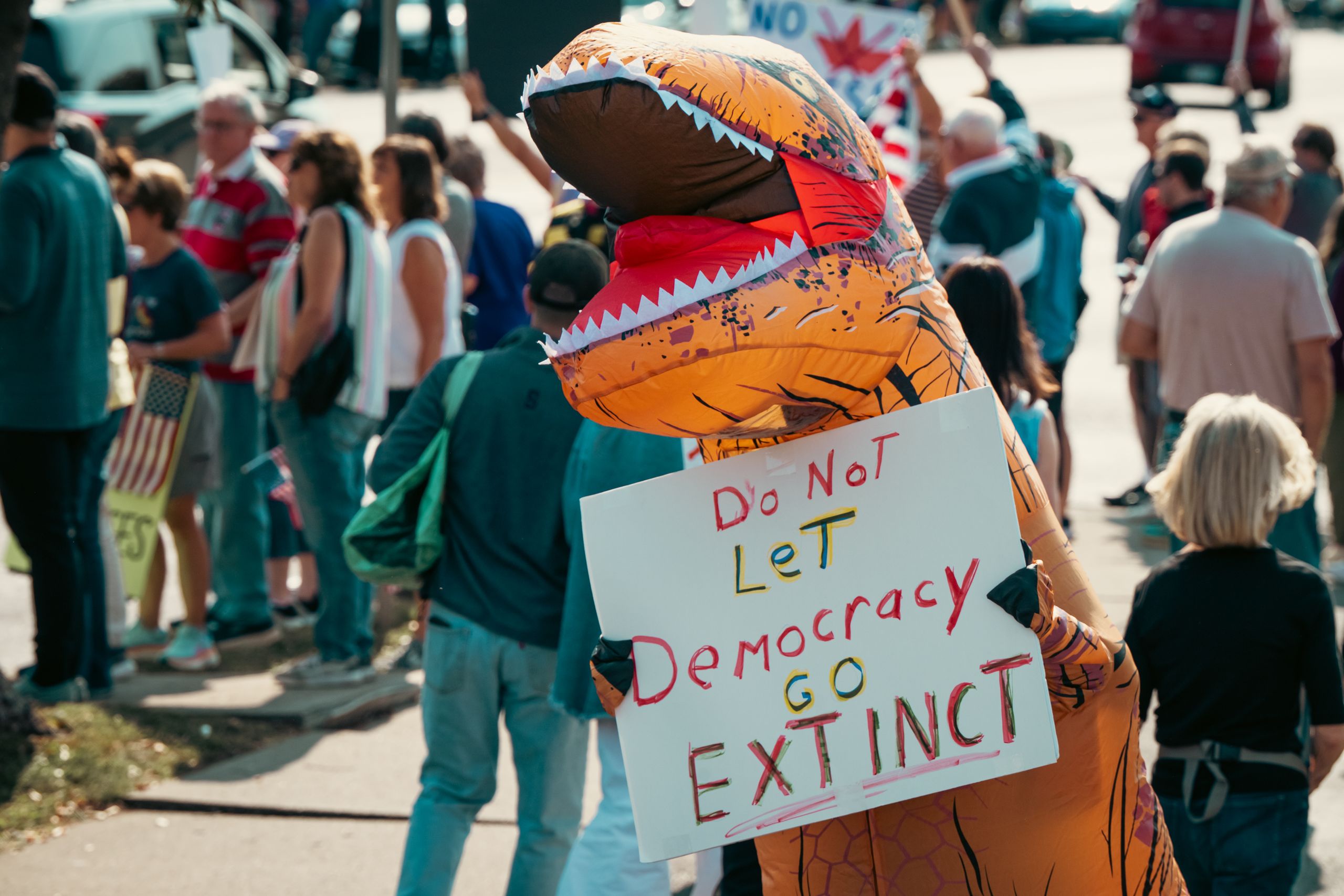 A person in a dinosaur costume holds a sign advocating for democracy at a public protest.